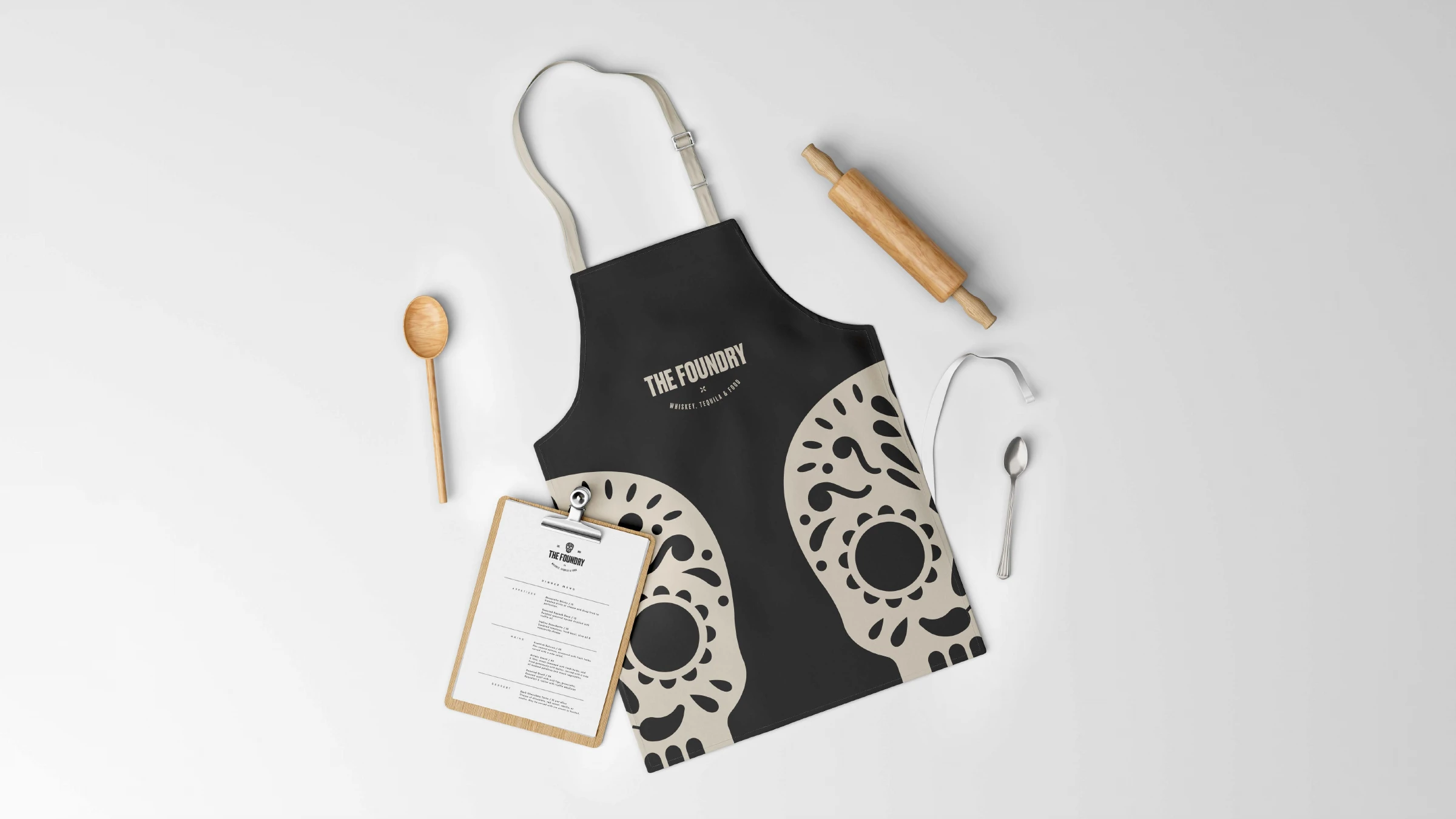 The Foundry staff apron with oversized skull motif, beside a branded menu clipboard